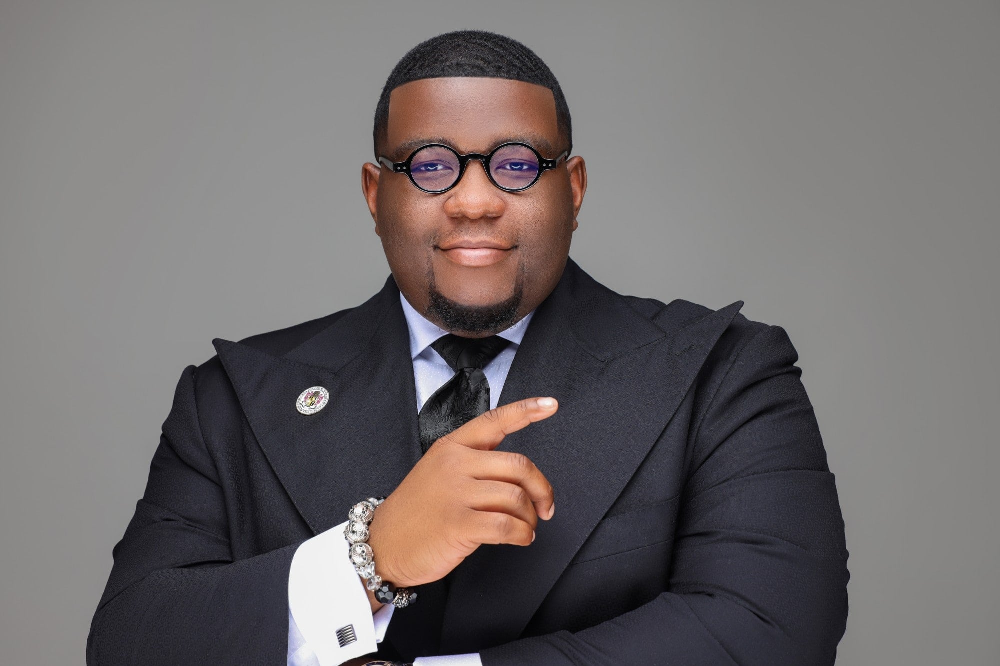 Overseer Jakeem Bowman in a black double-breasted suit and round glasses, smiling against a grey studio backdrop