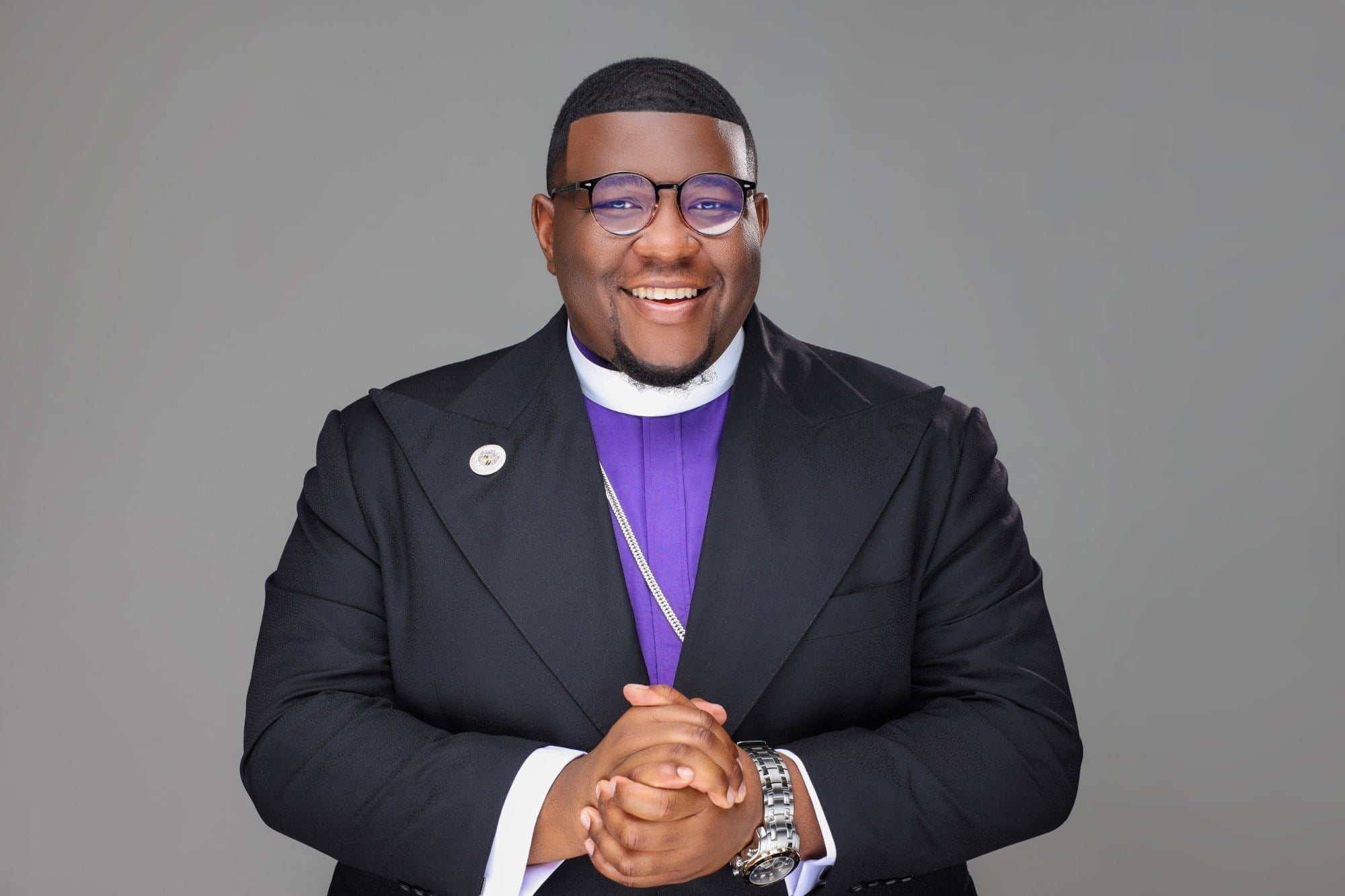 Overseer Jakeem Bowman smiling in a black suit over a purple clerical shirt and white collar, hands clasped